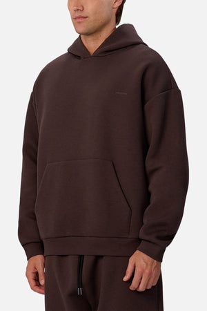 A man wears the Industrie The Tech Del Sur Hoodie in dark brown, featuring a loose fit, front pocket, and breathable fabric, paired with matching pants. He stands against a plain light background, showing only the upper half of his face.