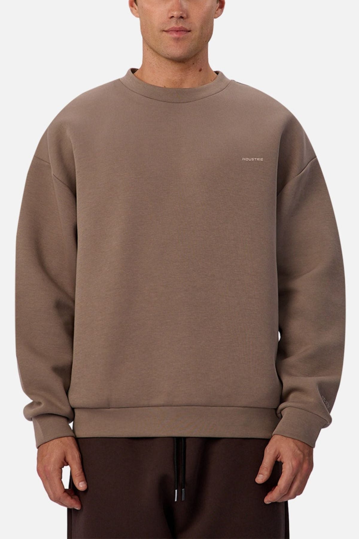 Someone wears the Industrie The Tech Del Sur Sweat— a loose-fitting, brown crewneck with long sleeves, athletic construction, and a small white logo on the upper left chest—paired with matching pants against a plain light background.