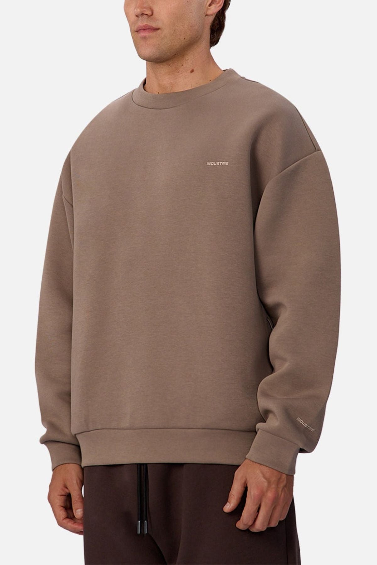 Someone stands against a plain white background wearing the Industrie The Tech Del Sur Sweat, a loose-fitting taupe top with small white text on the chest and sleeve, designed with athletic construction in mind.