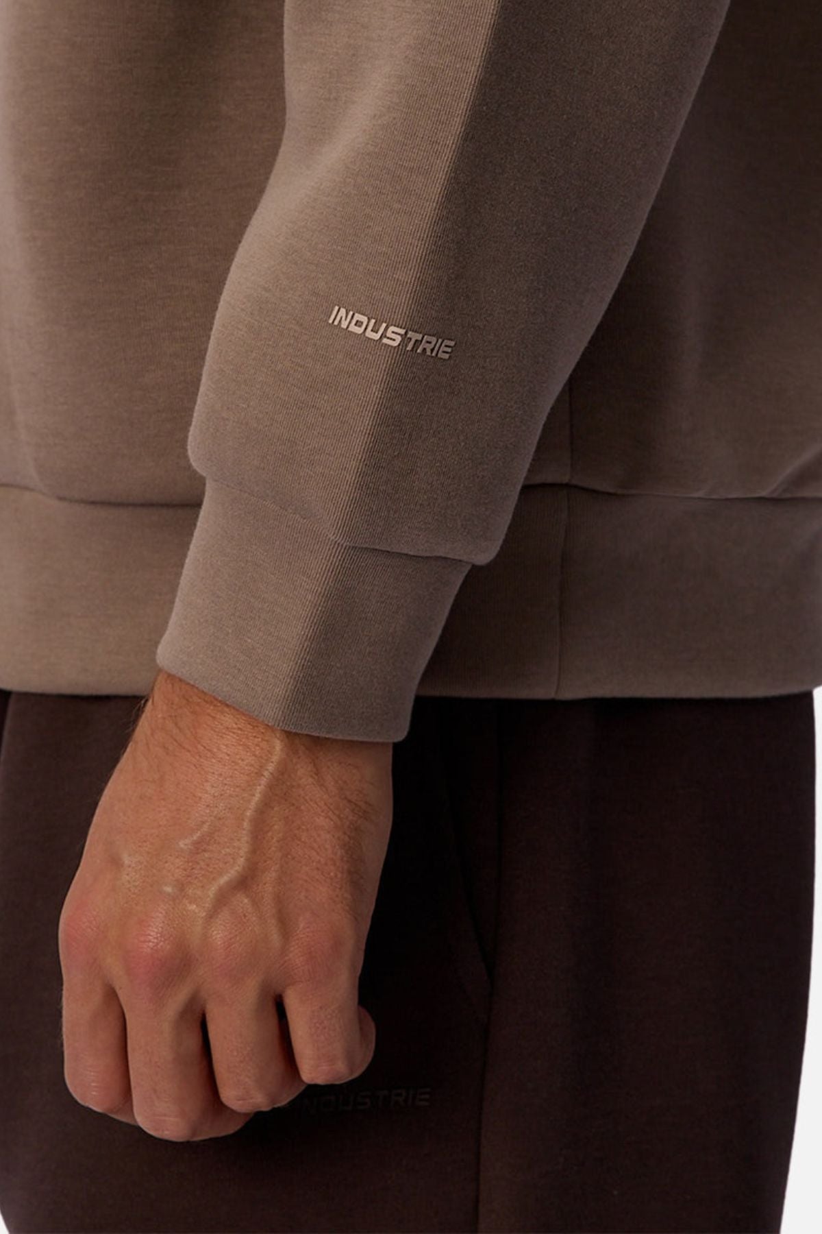 A close-up shows a person's arm and hand in the Industrie The Tech Del Sur Sweat, with "INDUSTRIE" on the taupe sleeve, paired with dark pants. Its athletic design suits active lifestyles. Set against a plain white background.