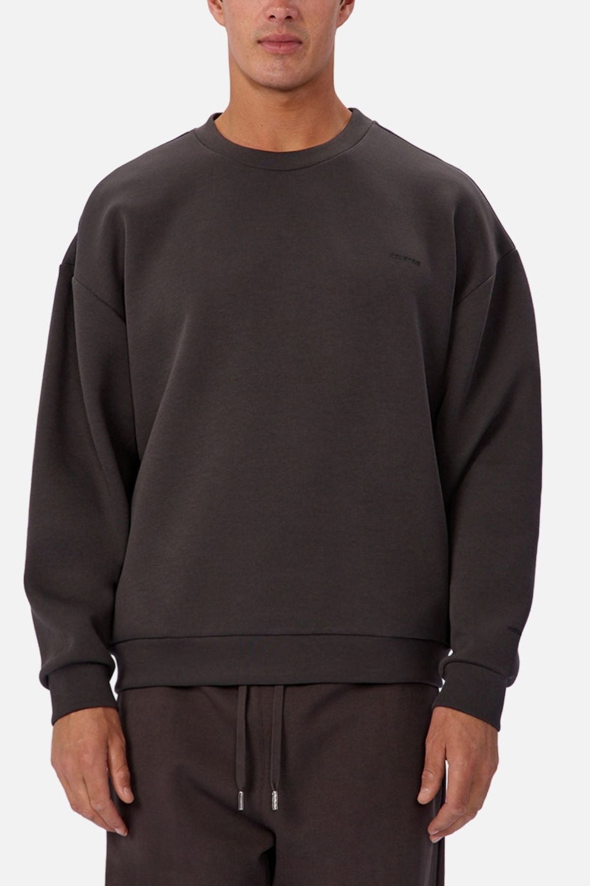 A person wears the Industrie The Tech Del Sur Sweat in loose-fitting dark brown, paired with matching drawstring pants, standing against a plain white background. Only the upper body and part of the legs are visible, suggesting an active lifestyle.