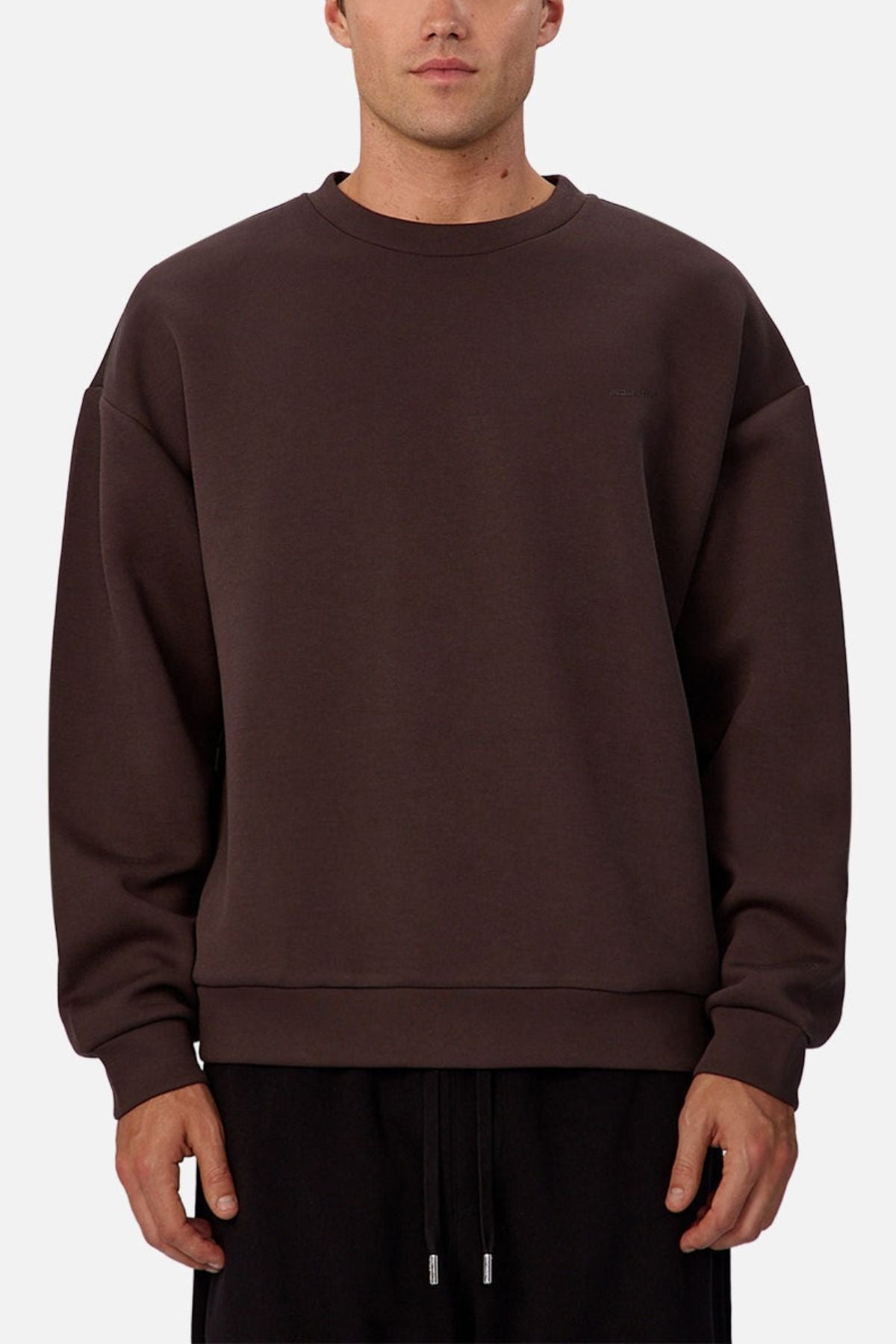 A man wears the Industrie The Tech Del Sur Sweat, a loose-fitting, dark brown crew neck with long sleeves, paired with black pants, standing against a plain light background.