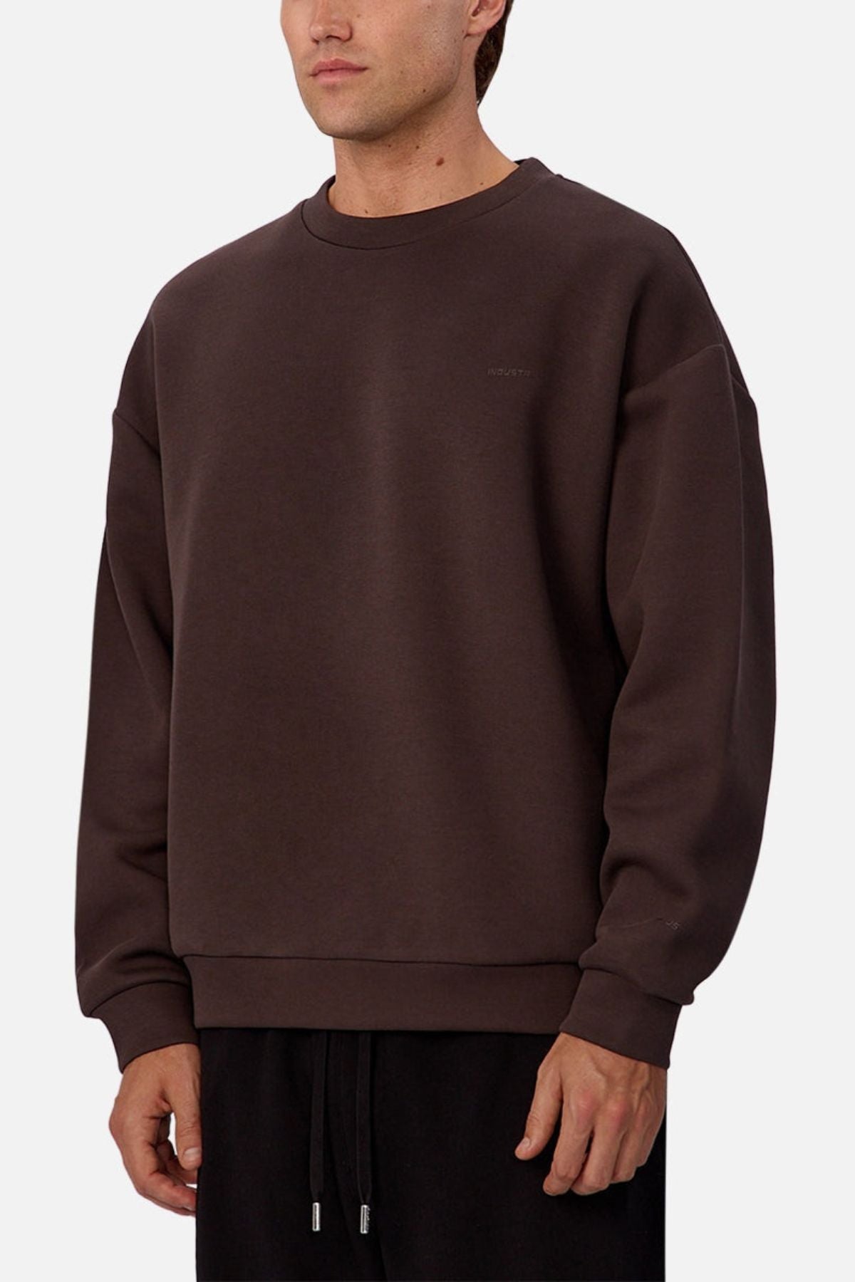 A person wearing a loose-fitting, dark brown Industrie The Tech Del Sur Sweat crewneck with long sleeves and matching jogger pants stands against a plain light background.