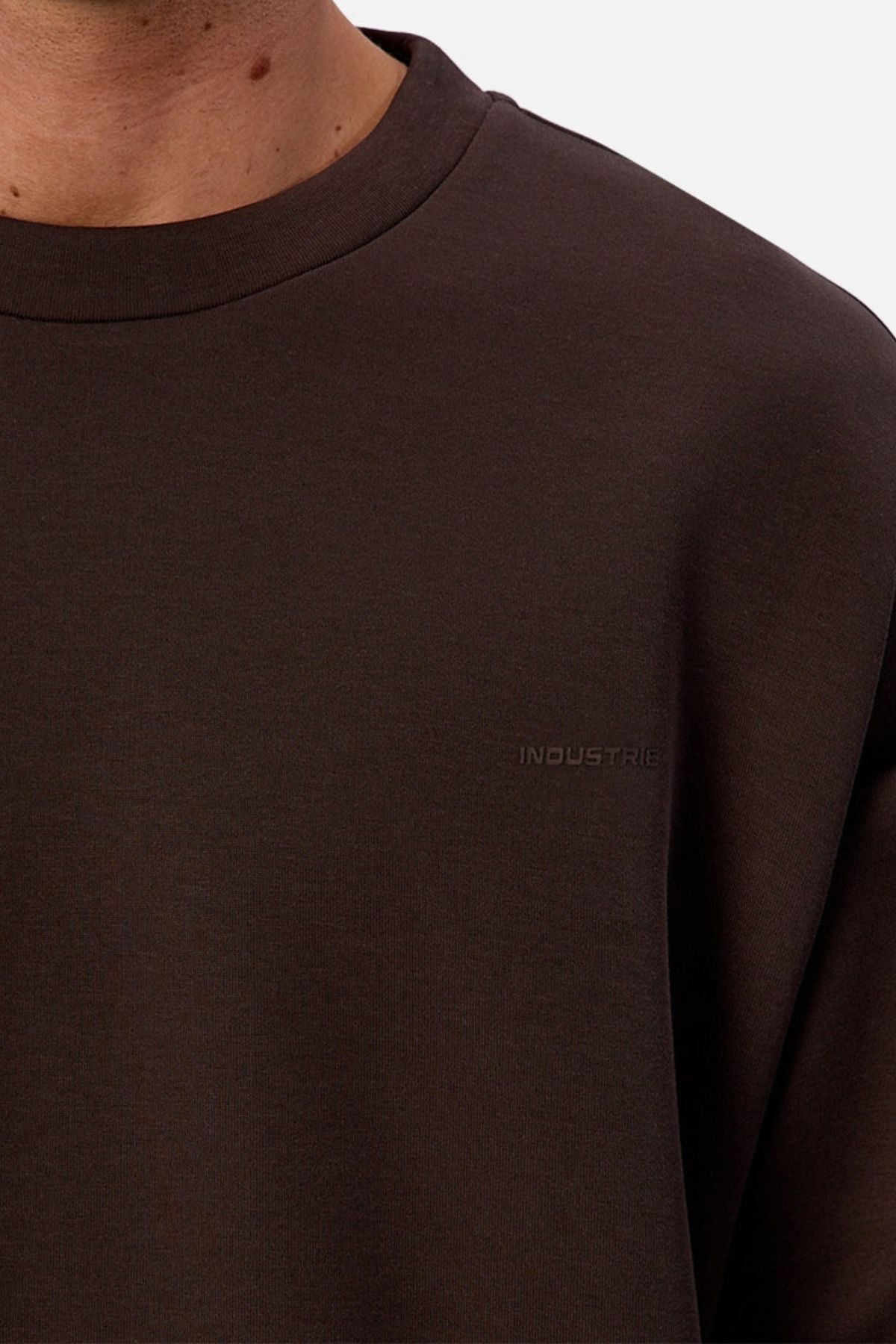 A close-up of a person wearing the dark brown Industrie The Tech Del Sur Sweat crew neck, featuring subtle "INDUSTRIE" embroidery on the chest. Only the lower face, neck, and upper chest are visible for a modern athletic style.