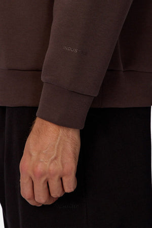 A close-up of a left hand and wrist wearing the Industrie The Tech Del Sur Sweat in brown with "INDUSTRIE" subtly embossed near the cuff, paired with black pants.