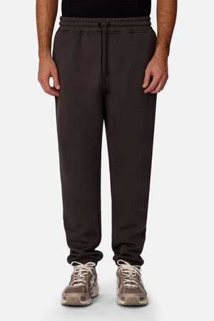 A person wearing Industrie The Tech Del Sur Track Pant in dark gray, made from athletic material, and light gray sneakers stands against a plain white background. Only the lower half of the body is visible.