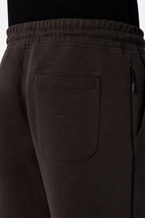 Close-up of the back pocket area of dark brown Industrie The Tech Del Sur Track Pant made from athletic material, featuring an elastic waistband and subtle "INDUSTRIE" embroidery on the pocket. The wearer pairs it with a black top.