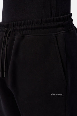 A close-up of a person wearing Industrie The Tech Del Sur Track Pant in black, featuring an elastic waistband with a drawstring and a small "INDUSTRIE" logo printed near the right pocket.