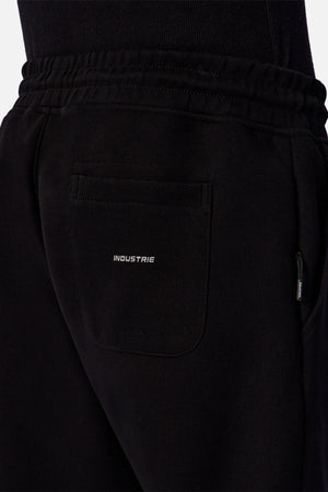Close-up of a person wearing black Industrie The Tech Del Sur Track Pant with an elastic waistband and a back pocket displaying the white "INDUSTRIE" logo, paired with a black top.