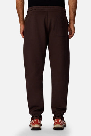 A person is seen from behind wearing Industrie The Tech Del Sur Track Pant in dark brown with a rear pocket, made from athletic material, paired with red athletic shoes featuring white soles. The background is plain and light-colored.
