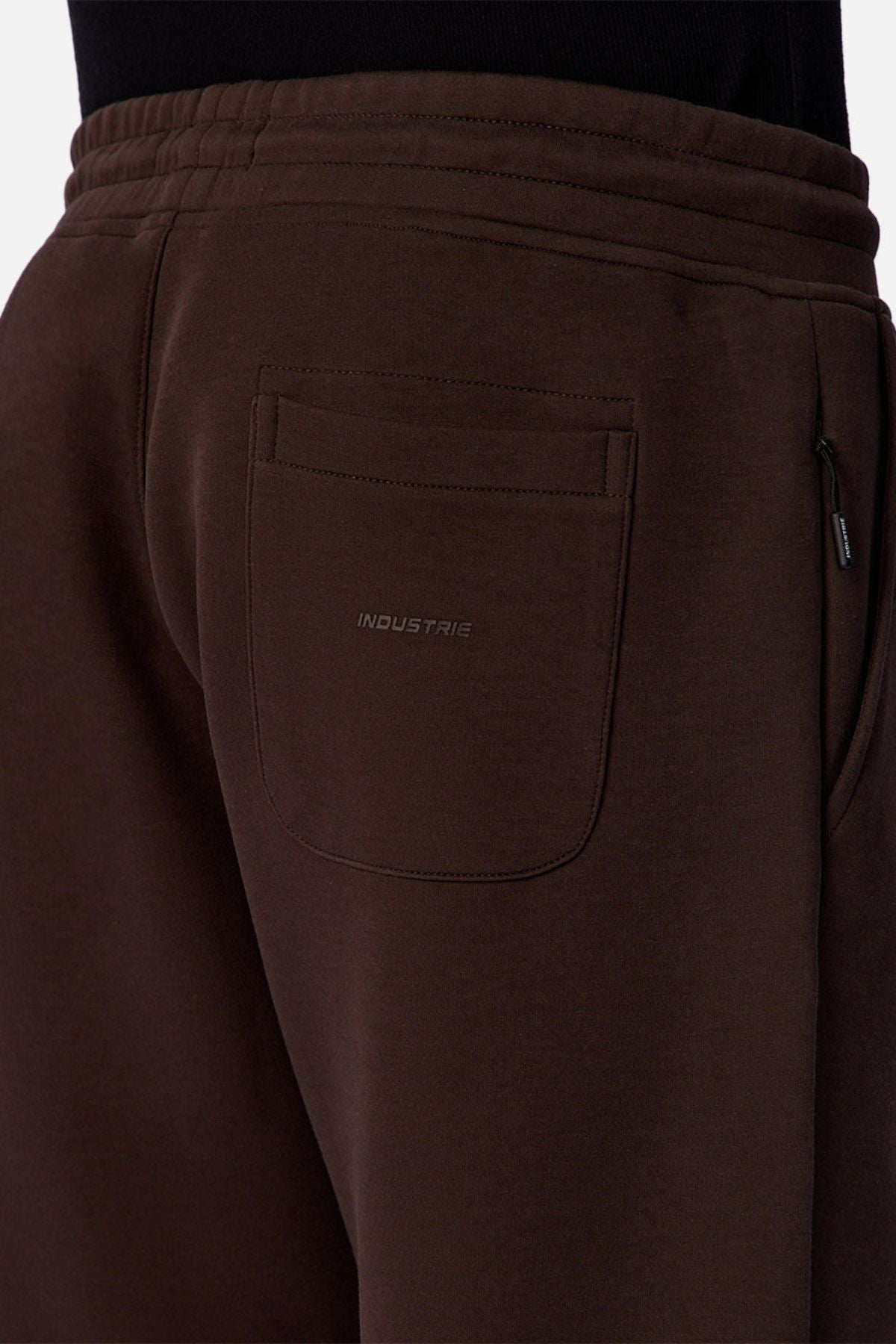 Close-up of a person wearing the Industrie The Tech Del Sur Track Pant in dark brown, featuring embroidered branding above the back pocket. The athletic material, waistband, and side pocket are partially visible.
