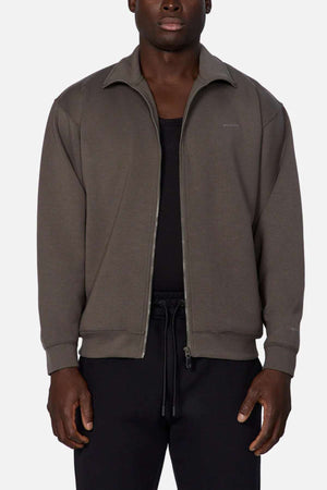 A man wears the Industrie The Zip Thru Track Top, made from soft dark gray fabric in an athletic style, layered over a black shirt and black pants, standing against a plain light background.