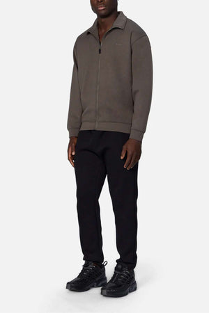 A man faces forward wearing the Industrie The Zip Thru Track Top in dark grey, made from soft, trendy fabric, paired with black pants and black athletic shoes against a plain white background.