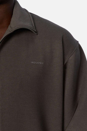 Close-up of someone wearing the Industrie The Zip Thru Track Top in dark grey, featuring soft fabric and "INDUSTRIE" embroidered on the chest. Only the shoulder and neck area show, emphasizing its athletic style.