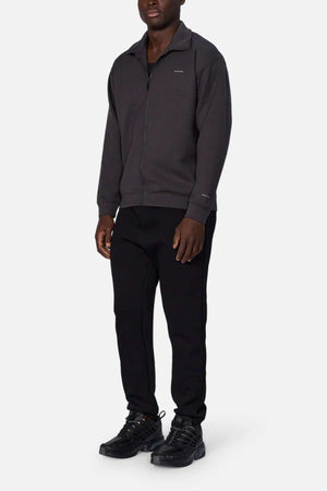 A person stands against a plain white background wearing the Industrie The Zip Thru Track Top, black pants, and black sneakers. The outfit is casual and monochromatic. The person has short hair and their hands rest by their sides, exuding an understated athletic style.