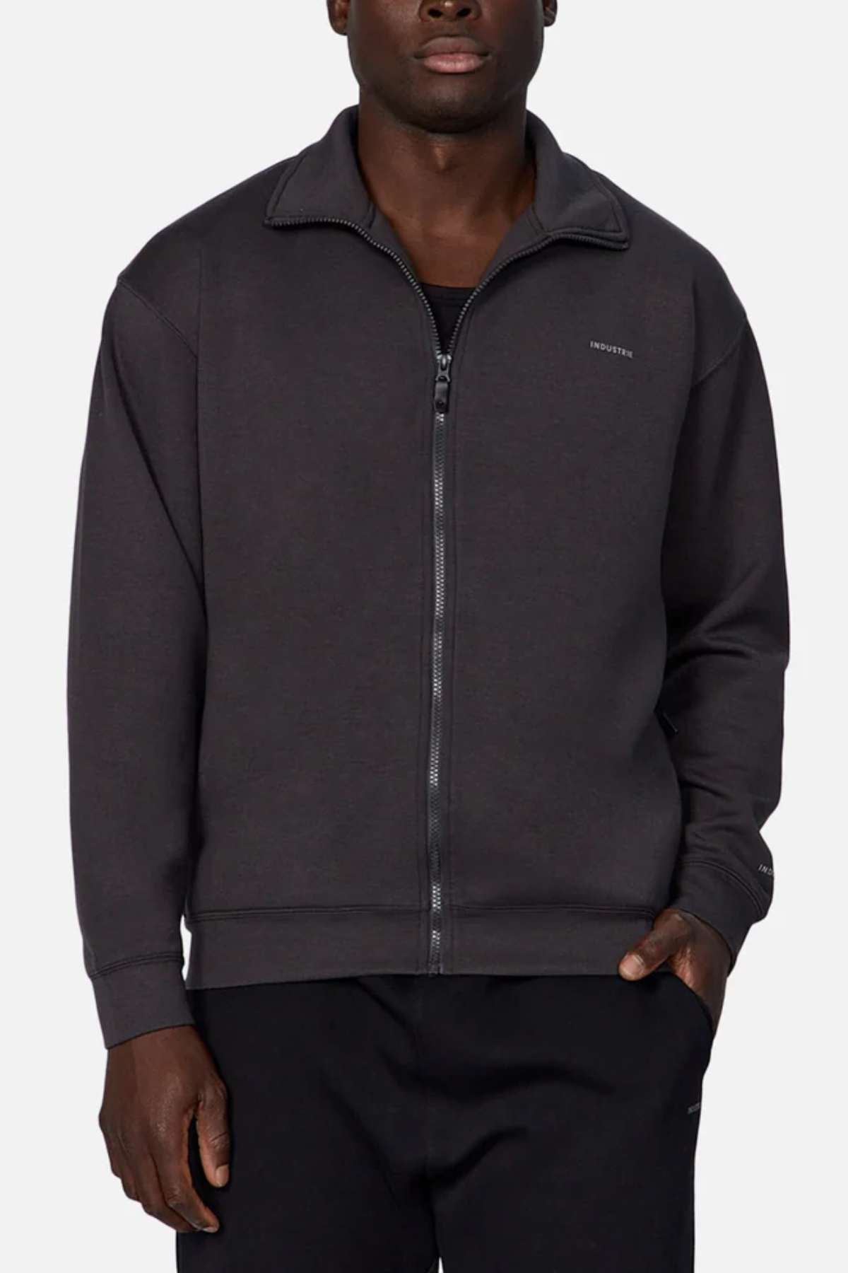 A person stands wearing the Industrie The Zip Thru Track Top in dark grey over a black shirt. The jacket, crafted from soft trendy fabric, features a high collar and subtle shoulder stitching. With their right hand in their pocket and left arm hanging by their side, they exude an athletic style against the plain white background.