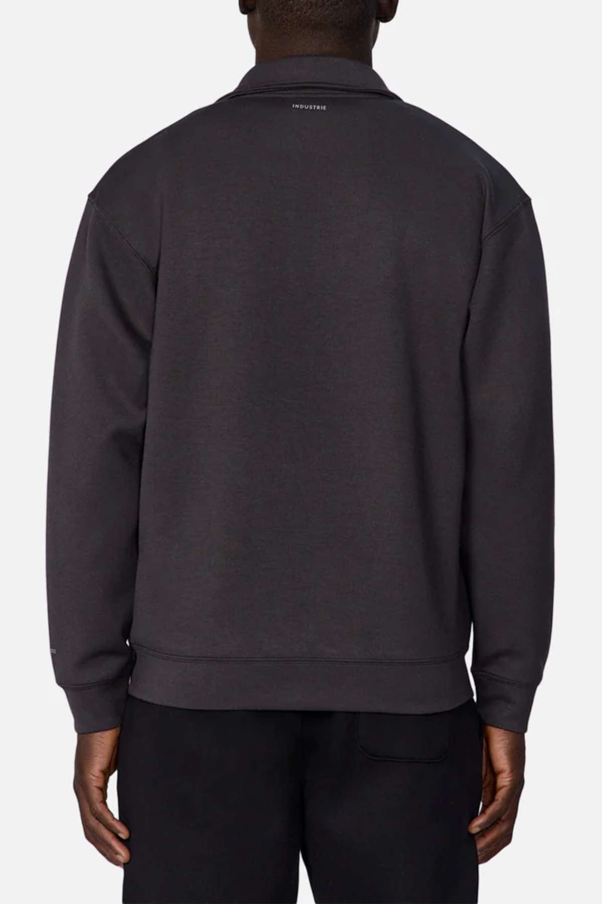 A person is shown from the back wearing Industrie The Zip Thru Track Top in dark gray and black pants. The soft, trendy fabric of the track top features a small text logo near the neckline and offers a relaxed fit. The plain white background highlights its understated athletic style.