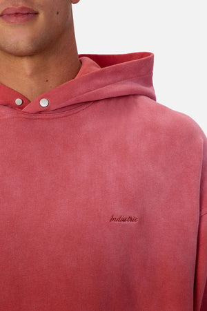 Someone wears the Industrie The Sandblast Hoodie in faded red, featuring silver snap buttons and "Industrie" embroidery on the chest. Made from 100% cotton, only their lower face and upper torso are visible, highlighting its relaxed style.