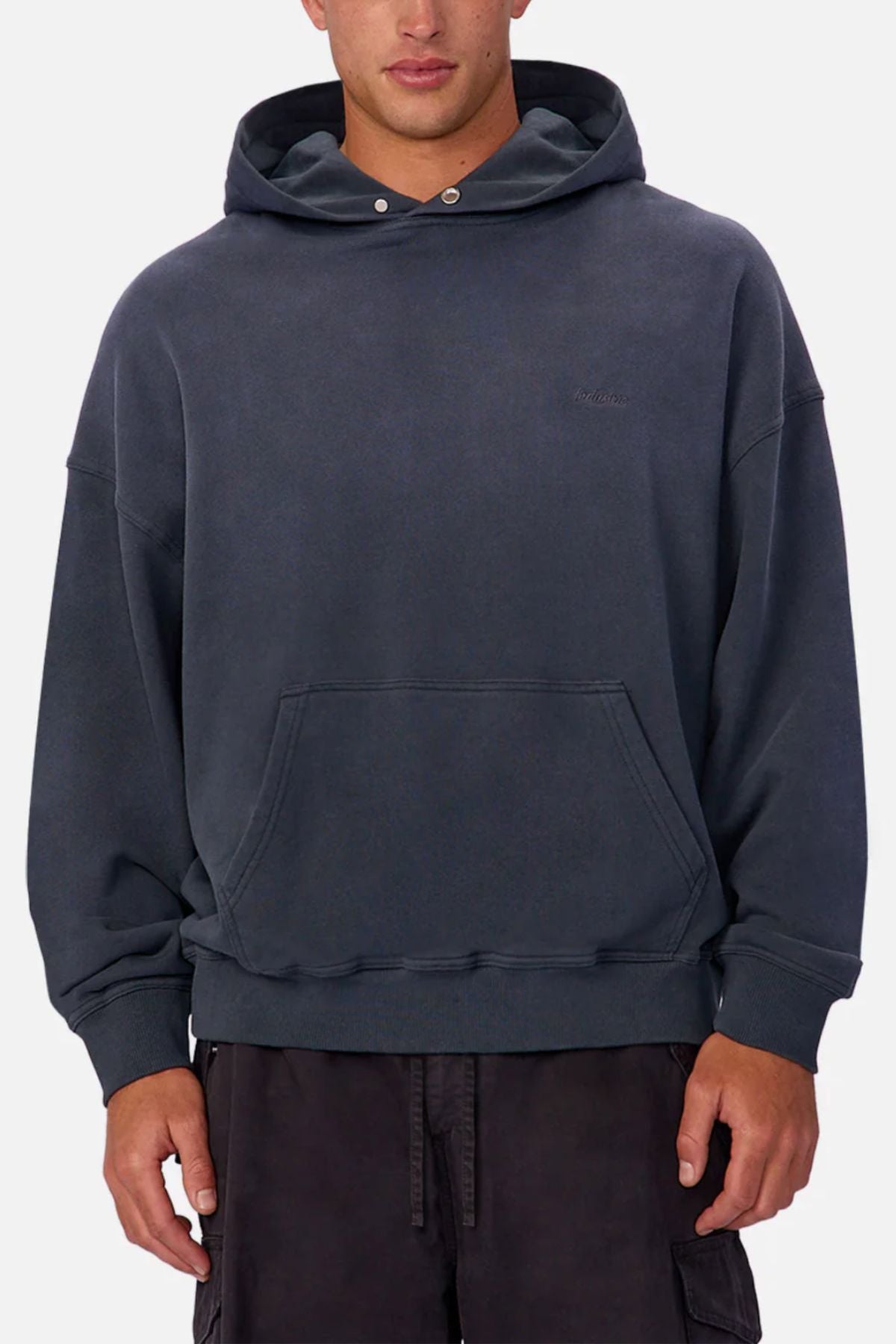 Someone in an Industrie The Sandblast Hoodie—loose-fitting, dark gray, 100% cotton with a subtle left-chest embroidered logo—pairs it with black pants against a light background for a relaxed look.