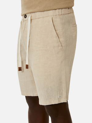 A person is donning beige Industrie The Baller Linen Shorts, featuring a drawstring waistband and a button, along with side pockets and a small brand label near the hemline. The knee-length shorts are matched with a beige top. The image centers on the individual's lower torso and upper legs against a plain white backdrop.