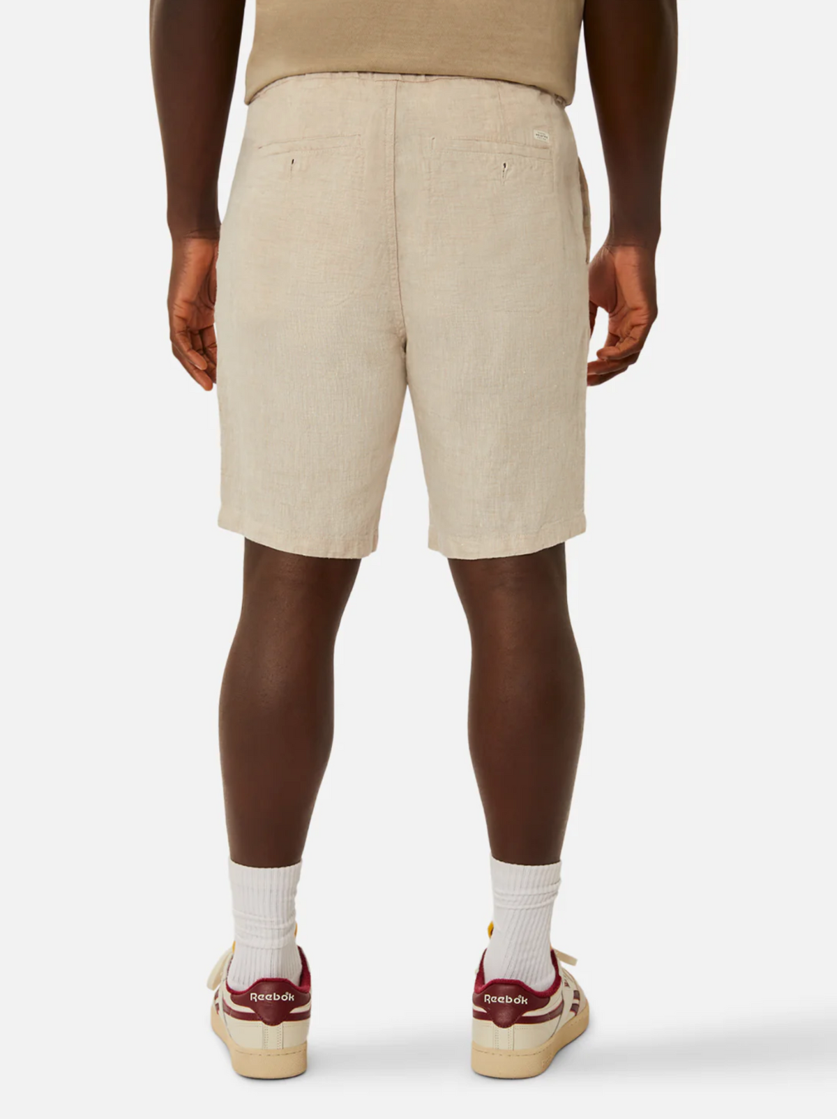 A person is standing with their back facing the camera, donning beige Industrie The Baller Linen Short with a drawstring waist, a beige top, white socks, and red-and-white Reebok sneakers. The plain white background ensures that the clothing and accessories remain the focal points of the image.