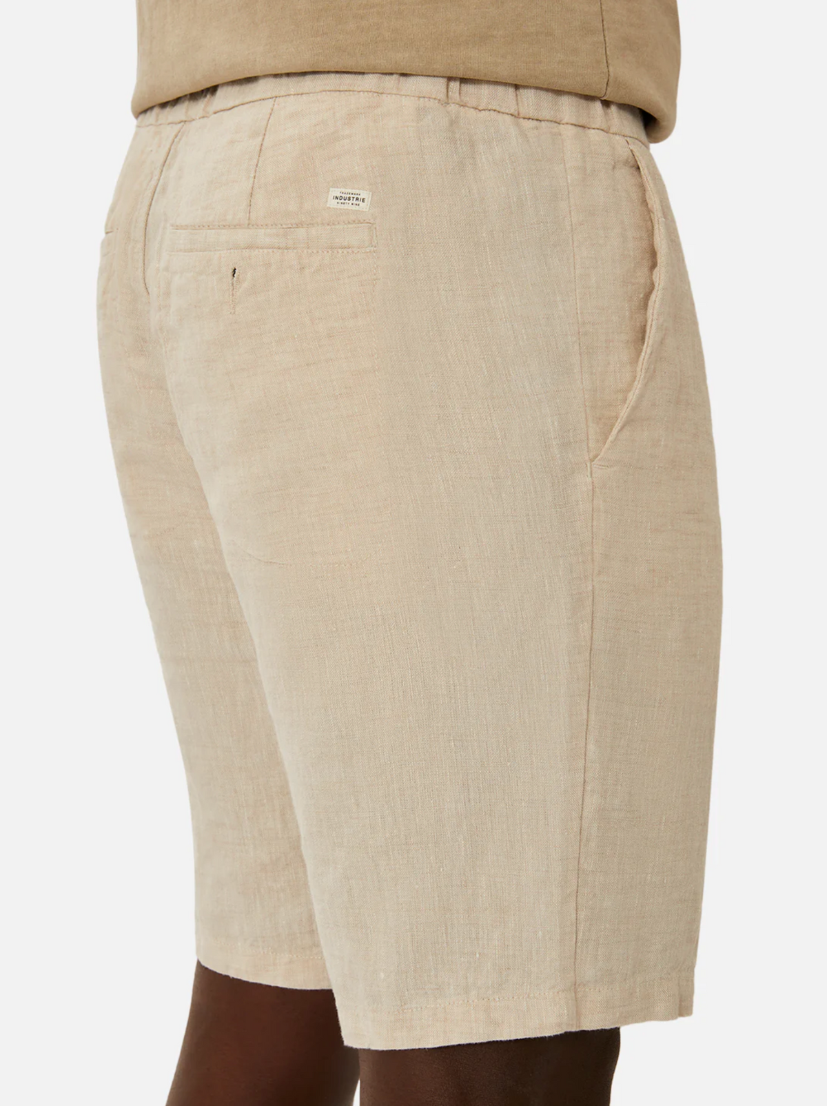 Close-up of the back and side view of a person's beige Industrie The Baller Linen Short. These knee-length shorts feature a back pocket with a small white label near the waistband and side pockets. The person is standing sideways with their hands out of view against a plain white background.