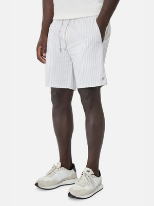 Wearing Industrie The Marchena Bahama Short with an elastic waist, a white shirt, and white athletic shoes with black soles, the person leans casually against a simple backdrop, perfectly capturing the relaxed summer vibe.
