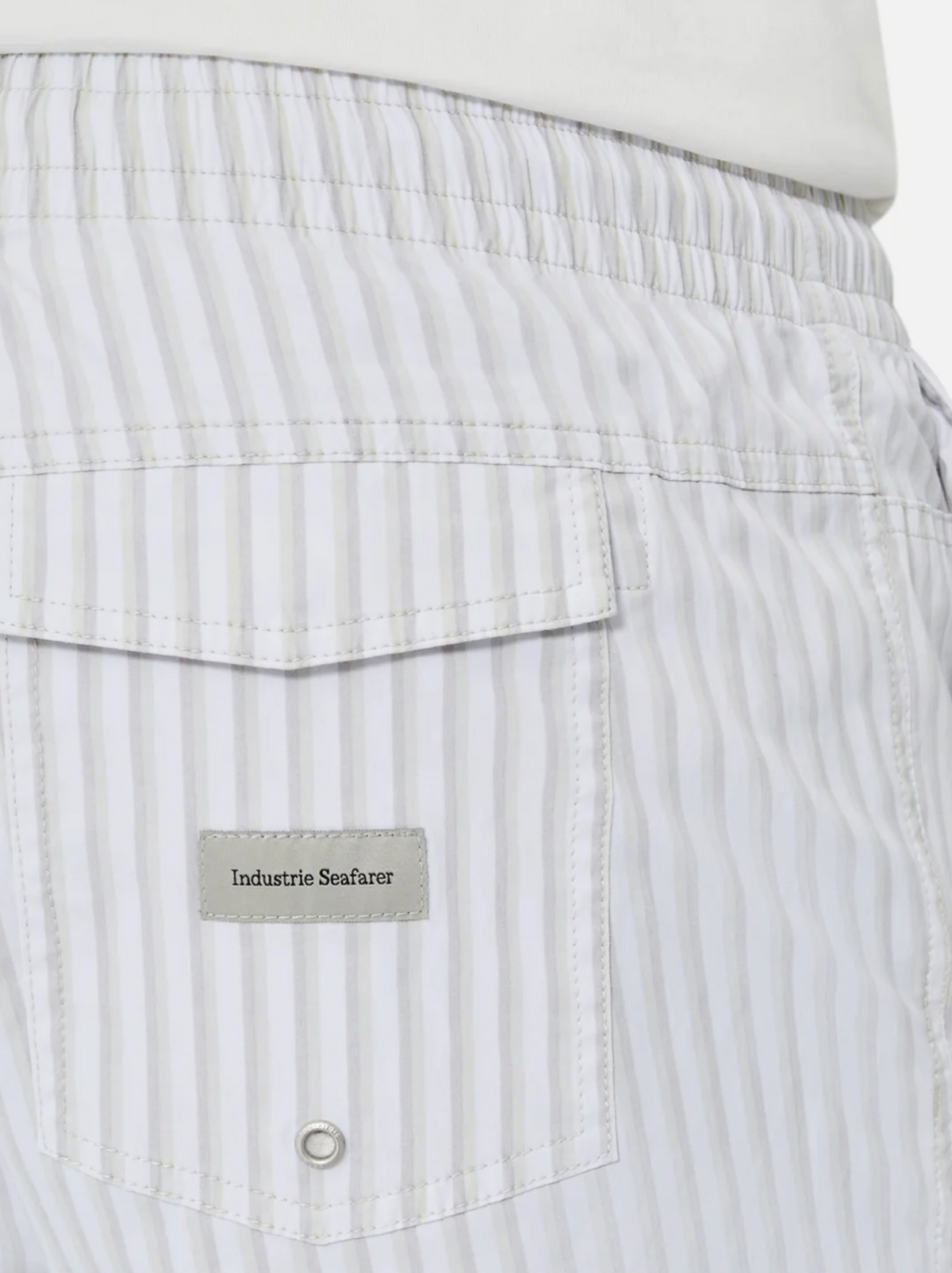 A person wears Industrie The Marchena Bahama Short featuring light gray and white vertical stripes with an elastic waist. Ideal for summer, it has a back pocket with an "Industrie Seafarer" label and a small metal eyelet below.