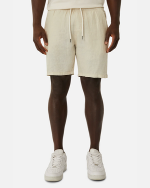 A person donning light beige Industrie The Marina Linen Shorts, crafted from pure linen with a drawcord waist, stands against a plain white background. Their hands are at their sides, revealing the waistband's drawstrings, perfectly paired with crisp white sneakers.