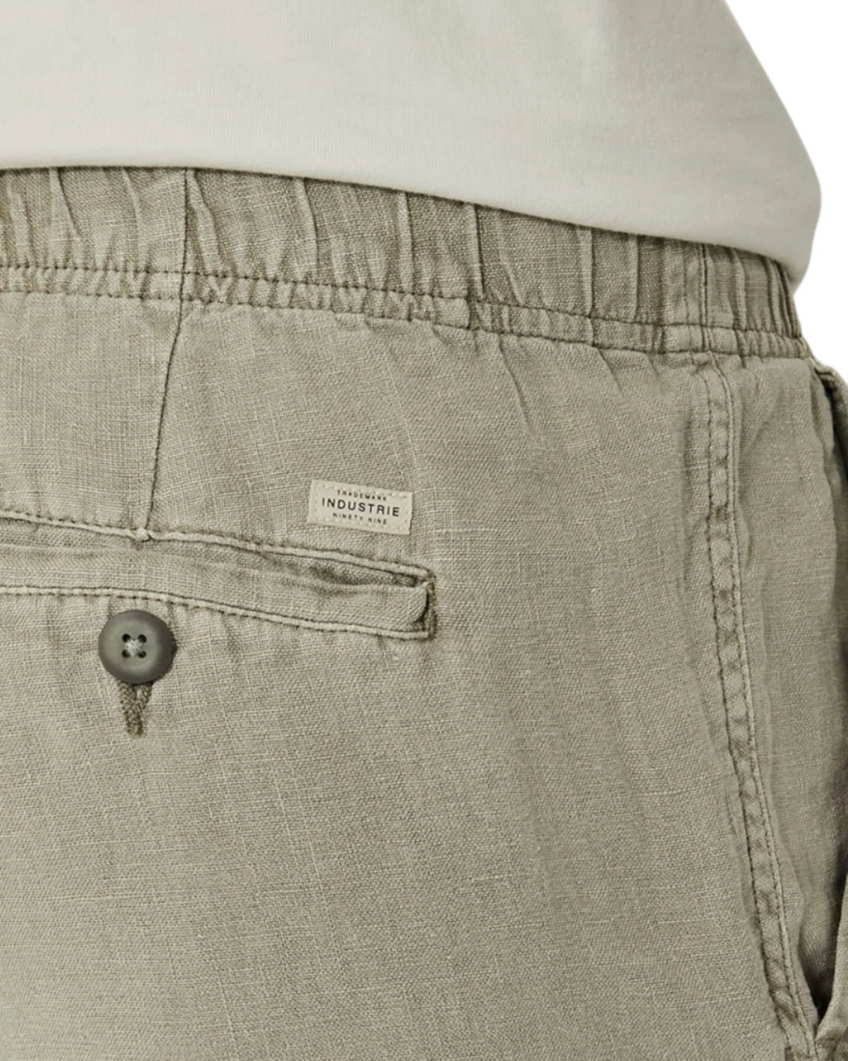 Close-up of the back pocket area of the Industrie The Marina Linen Short in light khaki, showing its elastic waistband, buttoned pocket with a small white "Industrie" label, and a white shirt peeking above the relaxed-fit waistband.
