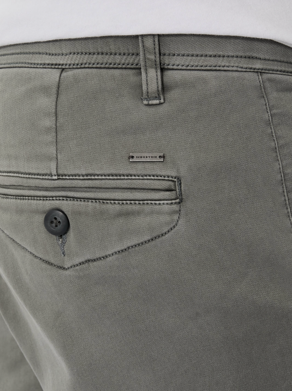 Close-up of the back pocket on the Industrie The New Drifter Cuba Short, a modern slim fit gray short with a black button and metallic brand label above the pocket. The waistband and part of a tucked-in white shirt are also visible.