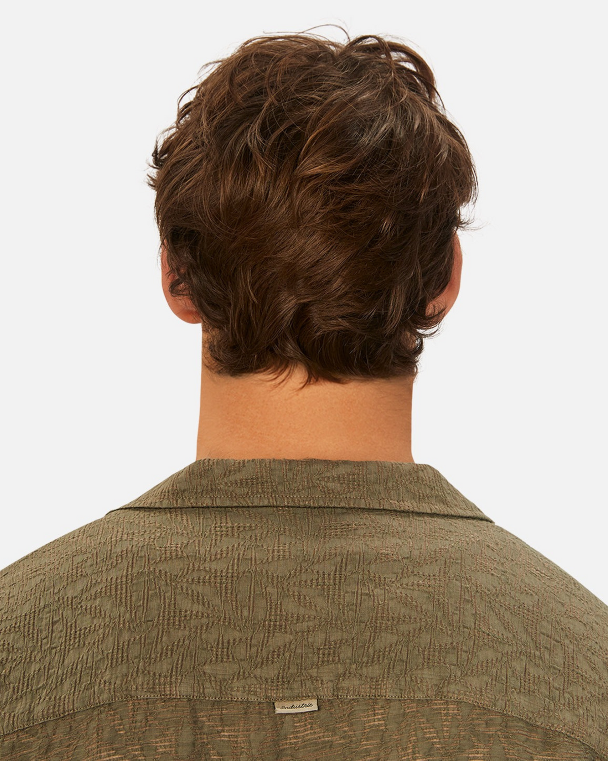The image features a person with wavy brown hair, seen from the back, wearing an olive green, loose-fit Industrie The Chiara Short Sleeve Shirt. Upper back and hair are highlighted, emphasizing the shirt's small tag at the spread collar against a plain white background.