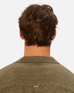 The image features a person with wavy brown hair, seen from the back, wearing an olive green, loose-fit Industrie The Chiara Short Sleeve Shirt. Upper back and hair are highlighted, emphasizing the shirt's small tag at the spread collar against a plain white background.