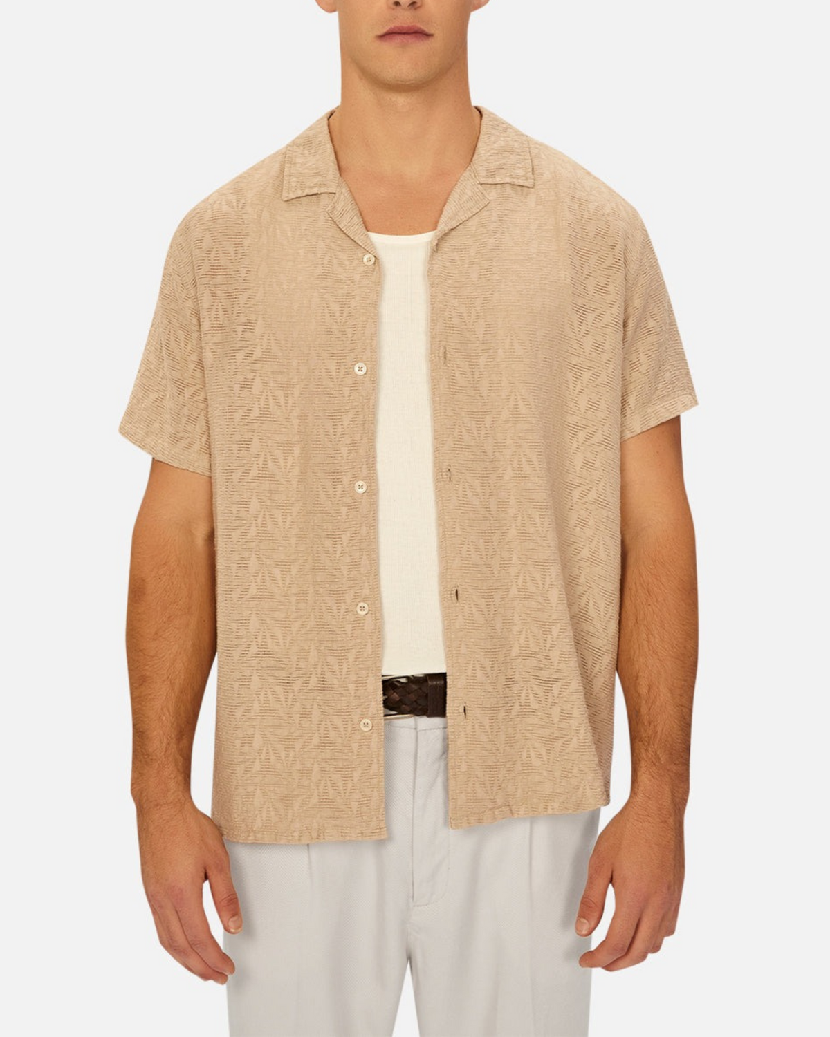 A man stands against a plain background wearing the Industrie The Chiara Short Sleeve Shirt in beige with an all-over pattern over a white undershirt, paired with light gray pants and a brown belt.