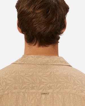 A person with short, wavy brown hair is seen from the back wearing the Industrie The Chiara Short Sleeve Shirt in beige, featuring an all-over pattern, notched collar, and loose fit.