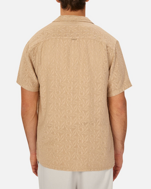 A man is seen from behind in a beige Industrie The Chiara Short Sleeve Shirt with all-over pattern, collar, subtle textured design, and loose fit. He pairs it with light pants against a plain white background.
