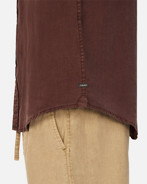 Close-up of a person wearing Industrie The Tennyson Linen Shirt in classic brown and beige pants. The 100% pure linen shirt features a small tag near the hem, focusing on textures and colors against a plain background.