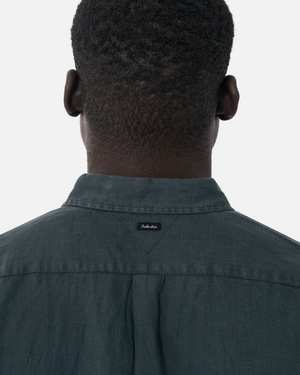 A person with short curly hair is seen from behind wearing the Industrie The Tennyson Linen Short Sleeve Shirt in dark green, featuring a small black label on the back collar. The background is plain white.
