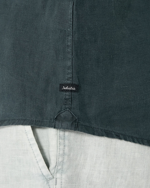 Close-up of the Industrie The Tennyson Linen Short Sleeve Shirt in dark green, featuring a center seam, short sleeves, and a small black "Industrie" tag above the hem, styled over light linen pants with a triangular stitch at the bottom edge.