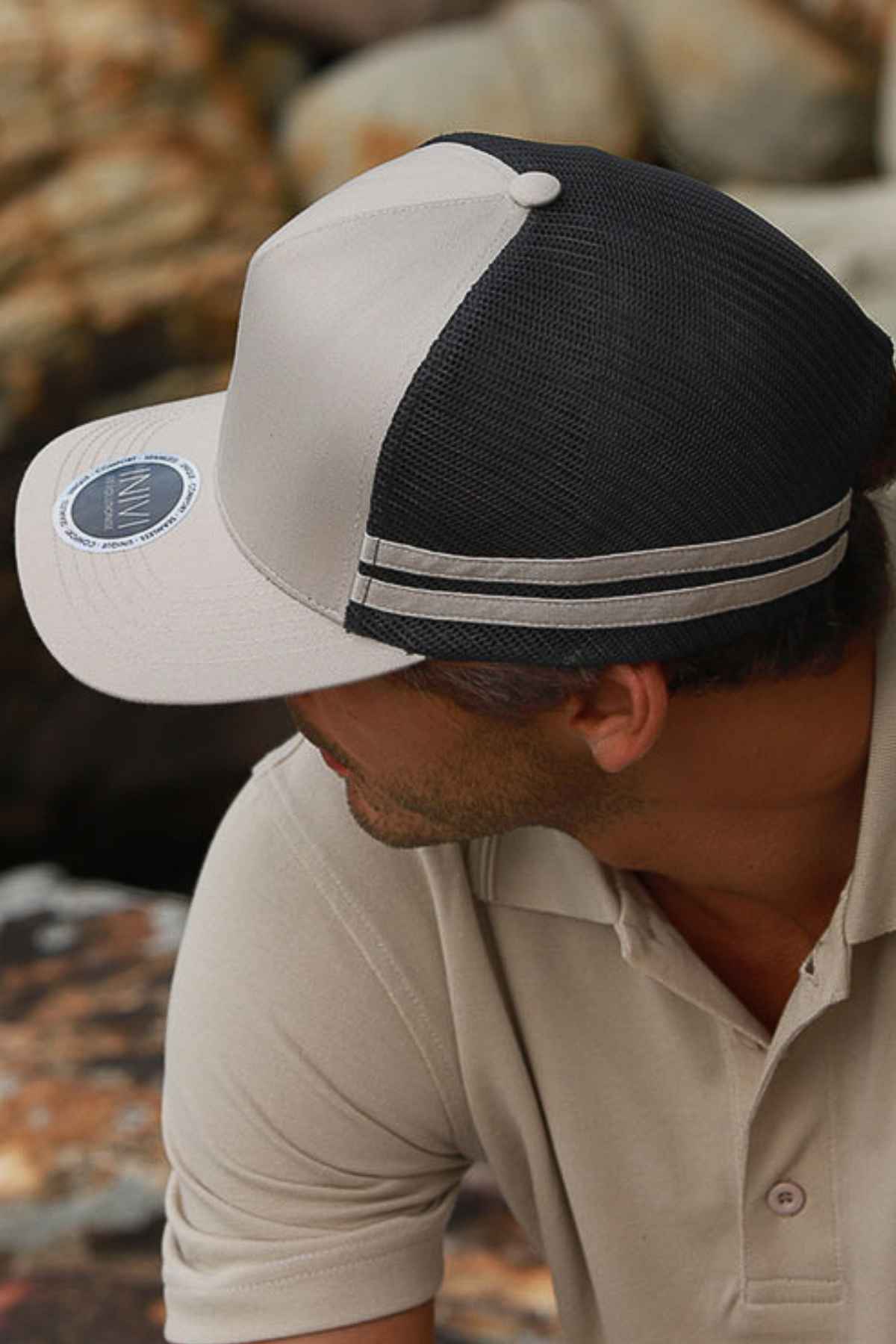 Wearing the Inivi Striped Trucker Cap in light gray and black with an adjustable strap, a person sits on a rocky surface. Dressed in a beige polo shirt and looking away, the focus is on the cap and shirt's comfortable and breathable everyday wear appeal, while the background is artfully blurred.