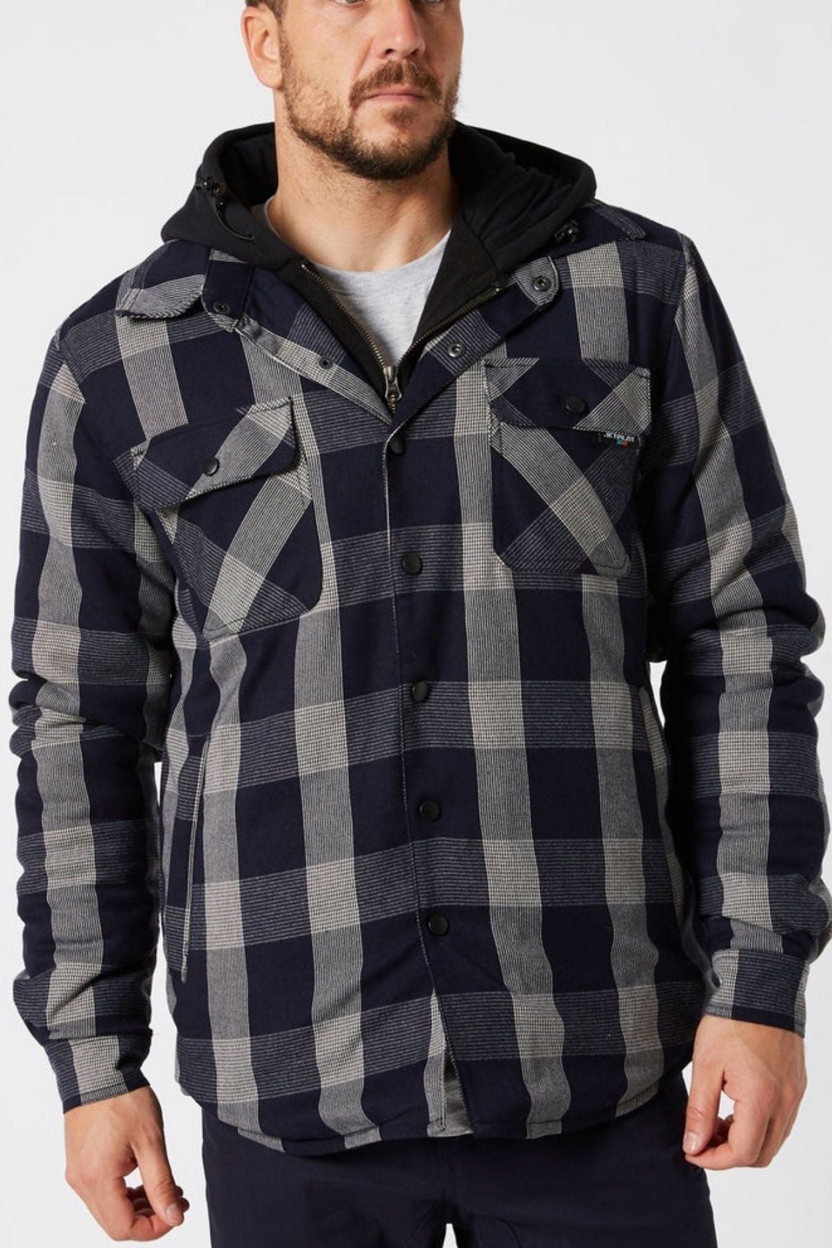 A man wears a Jetpilot Quilted Flannel Jacket in black and gray with front pockets, a black hood, and dark pants, standing against a plain white background.