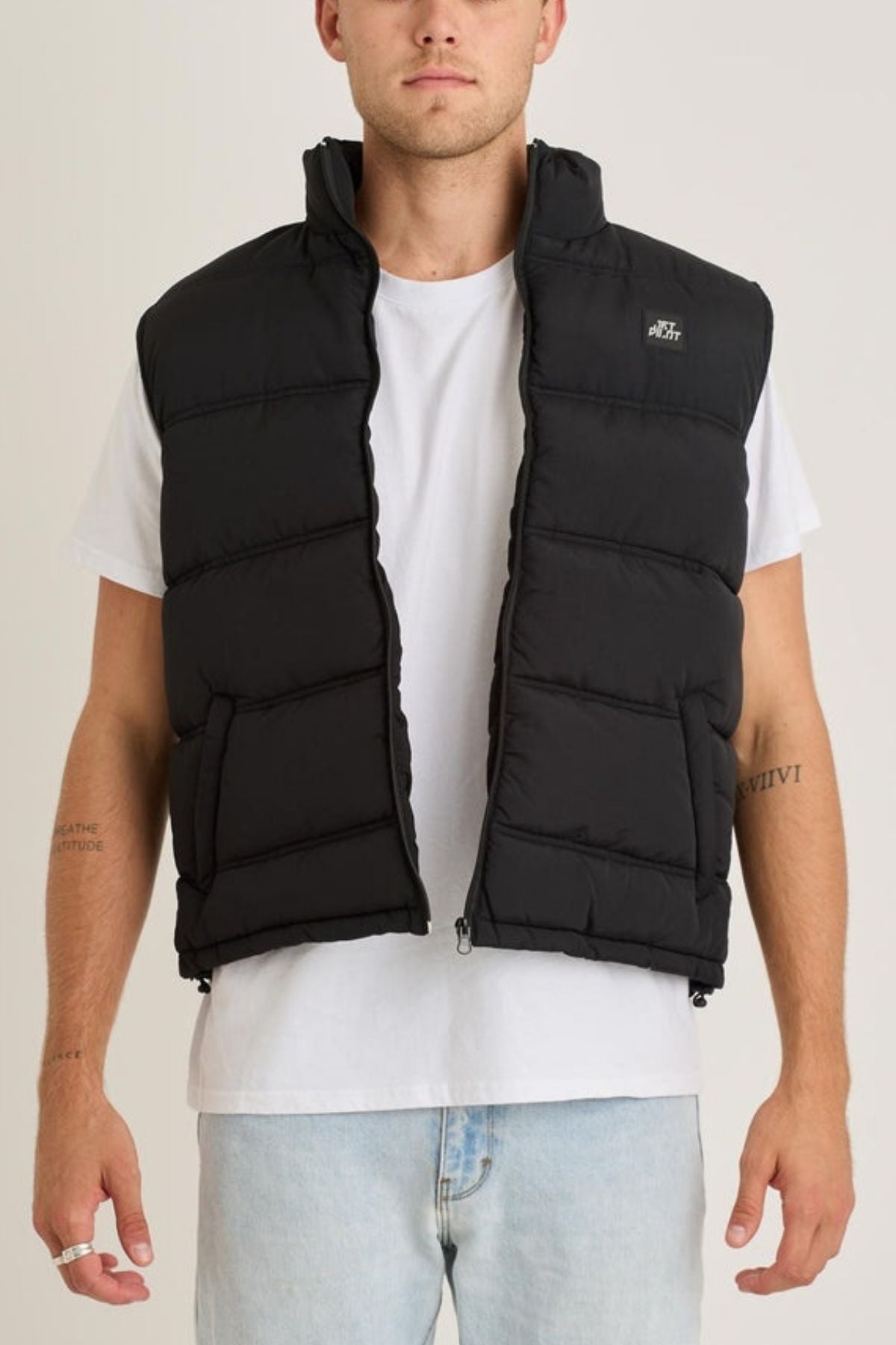 A person stands facing forward against a neutral background, wearing the Jetpilot Zip Up Vest over a white t-shirt and light blue jeans, with their hands relaxed at their sides.