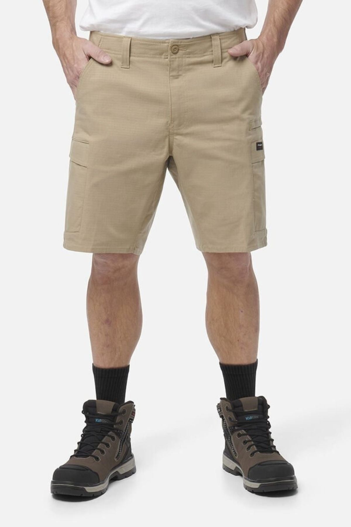 A person wears tan King Gee Workcool Pro Shorts made from durable Ripstop stretch fabric, paired with black socks and brown hiking boots, standing with hands in pockets against a plain white background. Only the lower body is shown.