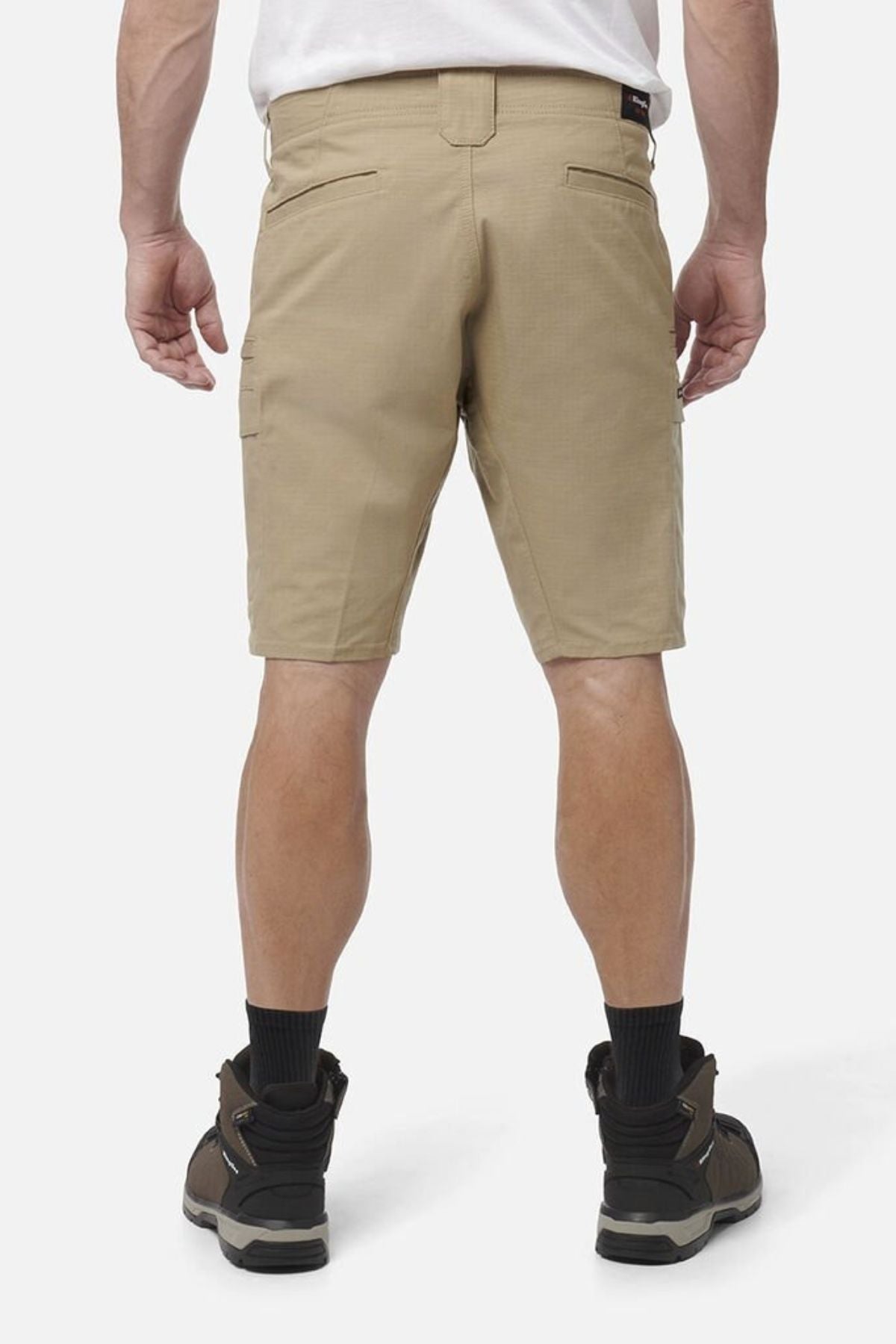 A person is shown from behind wearing a white shirt, black socks, brown hiking boots, and tan King Gee Workcool Pro Shorts made from ripstop stretch fabric, standing against a plain light background.