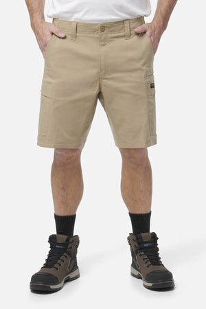 A person wears tan King Gee Workcool Pro Shorts made from durable Ripstop stretch fabric, paired with black socks and brown hiking boots, standing with hands in pockets against a plain white background. Only the lower body is shown.
