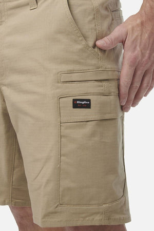 A close-up of a person wearing tan King Gee Workcool Pro Shorts made from ripstop stretch fabric, featuring side pockets and a KingGee logo patch above one pocket, with one hand resting by their side.