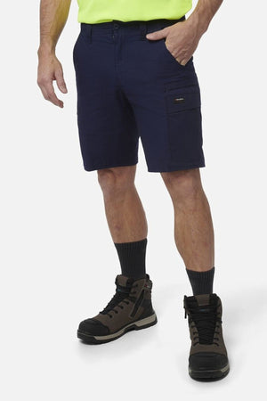 A person wearing navy King Gee Workcool Pro Shorts, a light yellow shirt, black crew socks, and brown work boots stands with one hand in their pocket against a plain white background.