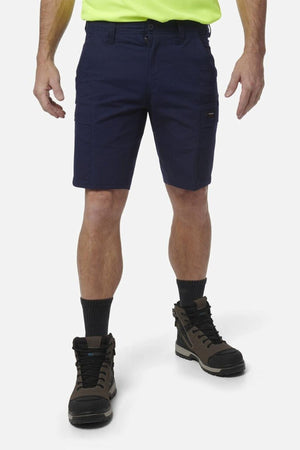 A person wearing navy blue King Gee Workcool Pro Shorts made from durable Ripstop stretch fabric, a neon yellow shirt, black socks, and brown work boots stands against a plain white background. The person's head is not visible in the image.