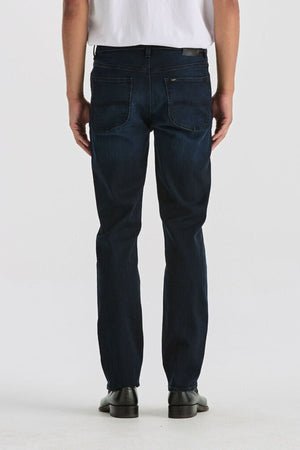A person in a white shirt, Lee Originals L100 Slim Jean with a comfortable fit, and black shoes stands facing away from the camera against a plain light background.
