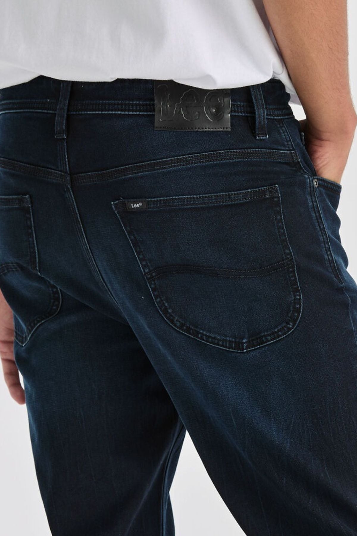 Close-up of a person wearing Lee Originals L100 Slim Jean in dark blue with a white shirt, hand in back pocket. The slim design ensures comfort and features a black leather "Lee" patch above the right pocket.